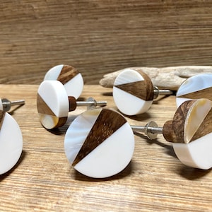 May include: A collection of round cabinet knobs with a modern design. Each knob features a white, wood, and shell inlay. The knobs have a silver-colored screw for installation. The wood has a dark brown color, and the shell has a pearlescent sheen.