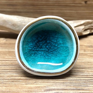 May include: A small, round ceramic bowl with a white exterior, a gold rim, and a turquoise interior. The interior has a crackle glaze effect. The bowl is likely handmade and has a unique, artistic appearance.