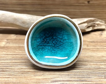 May include: A small, round ceramic bowl with a white exterior, a gold rim, and a turquoise interior. The interior has a crackle glaze effect. The bowl is likely handmade and has a unique, artistic appearance.