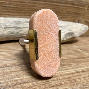 Könnte beinhalten: Ein ovaler Möbelknopf mit einer texturierten, hellorangen Steinoberfläche, die an den Seiten von zwei vertikalen, polierten goldfarbenen Metallbändern akzentuiert wird. Der Knopf hat einen silberfarbenen Metallstift an der Rückseite.