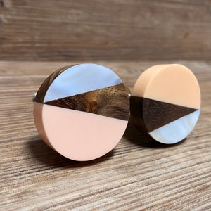 Pode incluir: Dois puxadores de armário redondos com um design geométrico. Cada puxador apresenta uma combinação de materiais: pêssego claro, madeira escura e branco perolado. Os puxadores estão sobre um fundo de madeira.