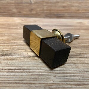 May include: A rectangular cabinet knob with a dark brown wooden body and a brass-colored metal accent. The knob has a screw for mounting and is designed for furniture. The wood appears to have a natural grain.