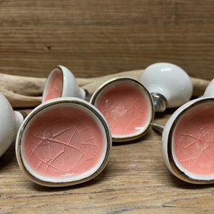 May include: A collection of ceramic cabinet knobs. Each knob features a white exterior, a cracked pink interior, and a gold-colored rim. The knobs are arranged on a wooden surface, suggesting a rustic or vintage aesthetic.