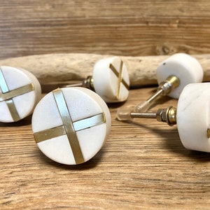 Könnte beinhalten: Eine Sammlung weißer Marmor-Schrankknöpfe mit Messingkreuz-Akzenten. Die Knöpfe sind rund und haben eine polierte Oberfläche. Sie werden auf einer Holzoberfläche präsentiert. Geeignet für Möbel und Heimdekoration.