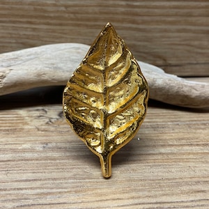 Może przedstawiać: Złoty, dekoracyjny przedmiot w kształcie liścia. Liść ma szczegółowe żyłkowanie i teksturowaną powierzchnię. Obiekt umieszczony jest na drewnianym tle z kawałkiem drewna wyrzuconego na brzeg.