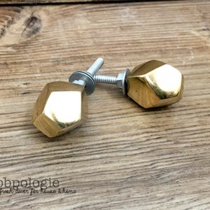 Peut inclure: Deux boutons de meuble dorés et facettés avec une quincaillerie argentée sont présentés sur une surface en bois. Les boutons ont un design géométrique, probablement pour des meubles. La marque "knobpologie" est visible.