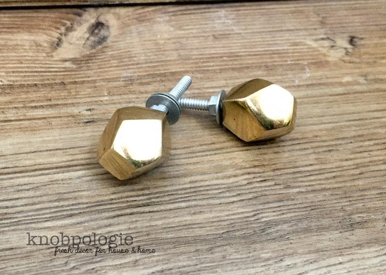 May include: Two faceted, gold-colored cabinet knobs with silver hardware are displayed on a wooden surface. The knobs have a geometric design and are likely for furniture. The brand name "knobpologie" is visible.