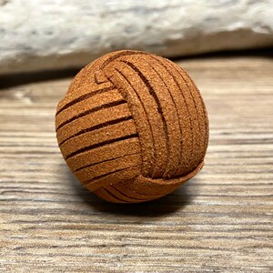 Op de afbeelding: Een klein, rond bruin leren object met een getextureerd oppervlak en horizontale snijlijnen. Het object lijkt op een miniatuurbal, mogelijk een decoratief element of een onderdeel van een groter item. Achtergrond is hout.