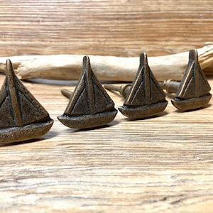 Könnte beinhalten: Vier bronzefarbene Schrankknöpfe in Segelbootform. Jeder Knopf hat ein detailliertes Segeldesign und einen kleinen Stiel zur Befestigung. Die Knöpfe sind auf einer Holzoberfläche angeordnet, was auf ein nautisches oder Küstenmotiv hindeutet.