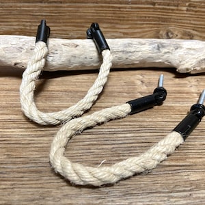 Peut inclure: Deux poignées en corde crème avec quincaillerie en métal noir. La corde est épaisse et torsadée, et les pièces métalliques ont une goupille argentée. Les poignées sont sur une surface en bois.