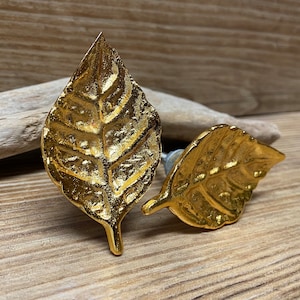 May include: Two gold-coloured leaf-shaped cabinet knobs. The knobs have detailed veining and a metallic finish. They are set against a wooden background, suggesting a nature-inspired or decorative theme.