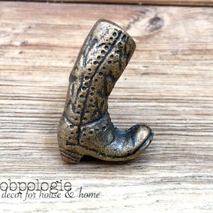 Peut inclure: Un bouton de meuble décoratif en forme de botte de couleur bronze. La botte présente des coutures détaillées et un design de botte de cowboy classique. Le texte "Knobpologie fresh decor for house & home" est visible en bas de l'image.