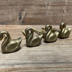 May include: Five gold swan-shaped drawer pulls. The pulls are made of metal and have a smooth, polished finish.