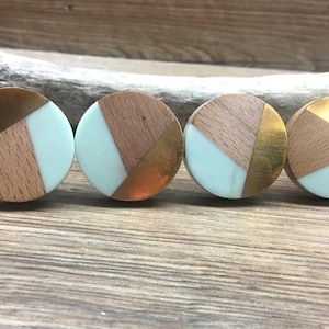 Puede incluir: Cuatro pomos redondos para armarios con un diseño único. Cada pomo presenta una combinación de madera, metal dorado y resina azul claro. Los pomos están dispuestos en fila, mostrando sus materiales y colores contrastantes.