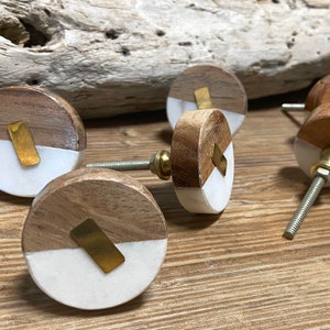 May include: A collection of wooden cabinet knobs with a two-tone design. The knobs feature a half-circle of light wood and a white section, accented with a brass-colored metal detail. The knobs are on a wooden surface.