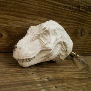 Op de afbeelding: Witte kastknop in de vorm van een dinosaurushoofd met een messing steel. De knop heeft een getextureerde afwerking en is geplaatst tegen een houten achtergrond. De tekst "knobpologie" is zichtbaar.