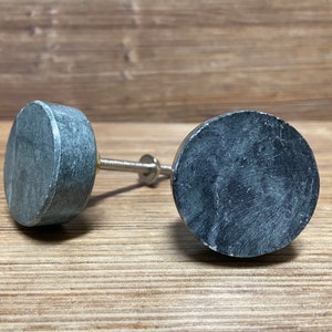 May include: Two circular cabinet knobs featuring a dark gray, mottled stone-like texture. Each knob has a flat, round face and a cylindrical base, connected to a threaded metal bolt with a small washer and nut for installation purposes.