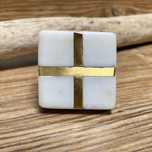 May include: A square, white marble cabinet knob with a brass cross design. The brass accents create a striking contrast against the white marble. This decorative hardware adds a touch of elegance to any furniture piece.