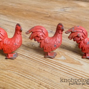 Puede incluir: Tres tiradores de armario rojos con forma de gallo y detalles envejecidos. Los tiradores son de metal y tienen un aspecto rústico y vintage. Los tiradores de gallo están colocados sobre una superficie de madera, ideal para decoración rústica.