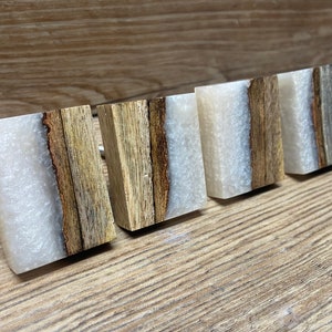 May include: Four square cabinet knobs featuring a combination of natural brown wood with a rough, textured edge and a smooth, translucent white resin section. Each knob has a distinct grain pattern and a rectangular shape with flat surfaces.