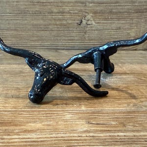 Puede incluir: Una cabeza de toro de metal negro con cuernos, con un tornillo para fijar. La cabeza de toro es un artículo decorativo, probablemente para muebles o decoración de paredes, con una estética rústica.