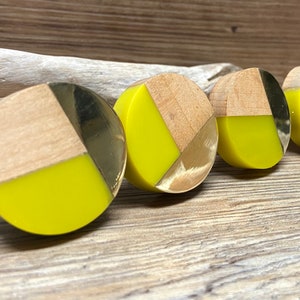 May include: Four circular cabinet knobs with a contemporary design. Each knob combines light wood, bright yellow, and metallic gold or silver accents. The knobs are arranged in a row on a wooden surface.