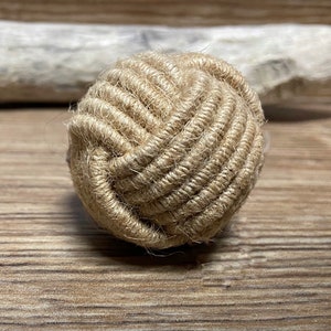 May include: A decorative, spherical knot made of natural-colored rope. The tightly woven rope forms a textured ball, showcasing a nautical or rustic aesthetic. The knot is displayed on a wooden surface.