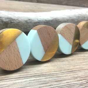 Puede incluir: Cuatro pomos redondos para armarios con un diseño geométrico. Cada pomo presenta una combinación de madera clara, metal dorado y una sección azul pálido. Los pomos están dispuestos en fila, mostrando su diseño único.