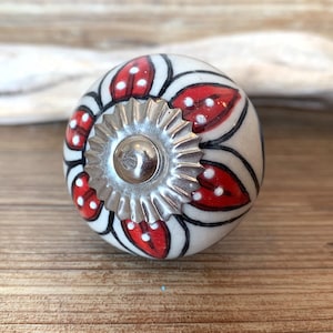 May include: A ceramic cabinet knob with a white base, adorned with red and white floral patterns and black outlines. The knob features a silver-colored metal base and a central screw. The design is reminiscent of a flower.