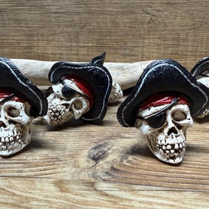 Puede incluir: Cuatro figuritas de calavera de pirata con sombreros de pirata negros y bandanas rojas. Cada calavera tiene un parche blanco sobre un ojo.