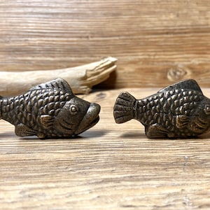 May include: Two antique gold-toned fish-shaped drawer pulls. The fish have detailed scales and a realistic appearance.