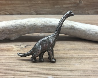 May include: A silver-colored metal dinosaur figurine in the shape of a brachiosaurus. The dinosaur is standing on a wooden surface.