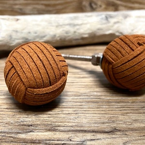 May include: Two brown leather cabinet knobs with a woven design. Each knob is spherical and attached to a metal screw. The knobs are set against a wooden background, suggesting a rustic or natural aesthetic.
