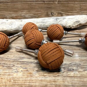 Peut inclure: Ensemble de sept boutons de meubles en cuir marron en forme de boule, avec des fixations argentées. Les boutons, disposés sur une surface en bois, présentent une texture distinctive. Idéaux pour personnaliser les meubles, chaque bouton mesure environ 4 cm de diamètre.