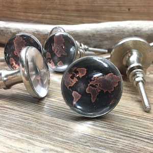 May include: A set of round cabinet knobs featuring a black and copper-coloured world map design under a clear glass dome. Each knob has a polished silver-tone metal base with a threaded bolt and nut for installation on furniture or cabinetry doors.