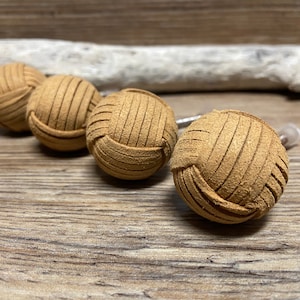 Peut inclure: Quatre boutons de meuble sphériques en cuir beige avec un motif cousu. Les boutons sont disposés en rangée, dont un légèrement flou. L'arrière-plan comprend une surface en bois et un morceau de bois flotté.