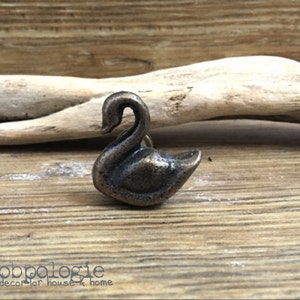 May include: A bronze-colored swan-shaped cabinet knob. The knob is detailed with a curved neck and a small, rounded body. The background features a wooden surface and a piece of driftwood. The text "Knobpologie fresh decor for house & home" is at the bottom.