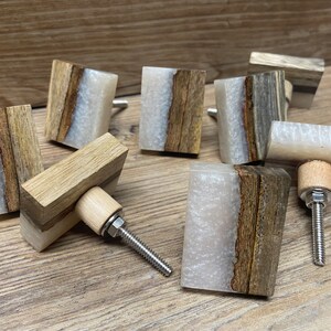 May include: A collection of square cabinet knobs featuring a combination of natural brown wood and translucent white resin. Each knob has a central wood section flanked by resin, with a metal bolt and a light-colored wooden base for mounting.