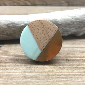 Puede incluir: Un pomo de armario redondo con un diseño en azul claro, madera y cobre. El pomo está dividido en tres secciones: una sección azul claro, una sección de madera y una sección de cobre. El pomo está sobre una superficie de madera.