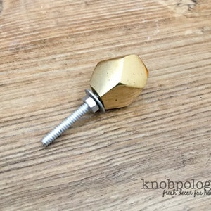 May include: A faceted, gold-colored cabinet knob with a threaded bolt, washer, and nut. The knob has a geometric design and is shown against a wooden surface. The text "knobpolgie fresh decor for house & home" is visible.