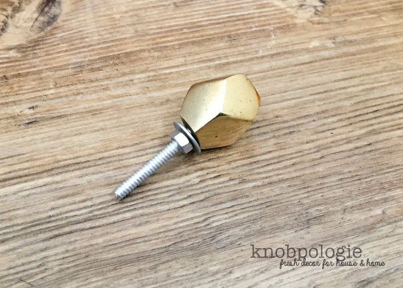 May include: A faceted, gold-colored cabinet knob with a threaded bolt, washer, and nut. The knob has a geometric design and is shown against a wooden surface. The text "knobpolgie fresh decor for house & home" is visible.