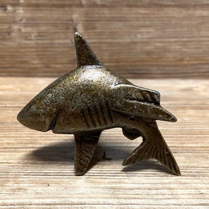 May include: A bronze-colored metal shark figurine. The shark is in a swimming position with its tail and fins extended.