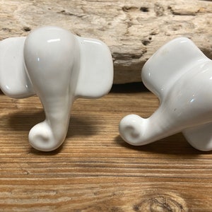May include: Two white ceramic elephant-shaped cabinet knobs. The knobs have a glossy finish and are designed with detailed trunks, ears, and heads. They are placed on a wooden surface.