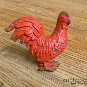Puede incluir: Un tirador de armario rojo con forma de gallo y acabado envejecido. La ferretería decorativa presenta un diseño detallado de un gallo con una superficie texturizada. El tirador es de metal y es adecuado para la decoración de cocinas o muebles.