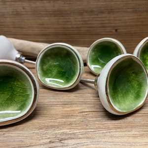 May include: A collection of six ceramic cabinet knobs. Each knob features a white exterior with a gold trim and a green, crackled interior. The knobs are mounted on silver hardware and rest on a wooden surface.