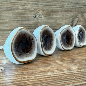 May include: Four geode-shaped cabinet knobs. Each knob has a white exterior with a gold trim and a dark brown interior. The knobs are arranged in a row on a wooden surface. The knobs are for furniture.