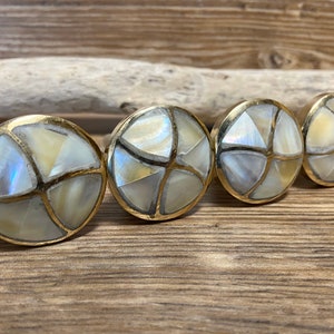 May include: Four round cabinet knobs with a mother-of-pearl inlay and gold-colored metal trim. The knobs are arranged in a row on a wooden surface, showcasing their iridescent, geometric design. These decorative knobs are ideal for furniture upgrades.