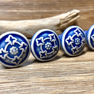 May include: Four blue and white ceramic cabinet knobs with a floral design. The knobs are round with a white rim and a blue centre featuring a white floral pattern. They are arranged in a row on a wooden surface.