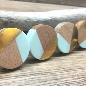 May include: Four cabinet knobs with a geometric design. Each knob features light wood, gold-coloured metal, and a section of pale blue. The knobs are arranged in a row on a wooden surface.