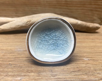 May include: A small, round ceramic bowl with a crackle glaze interior in shades of blue and white. The rim is trimmed with a gold band. The bowl is likely used for tea or small servings.
